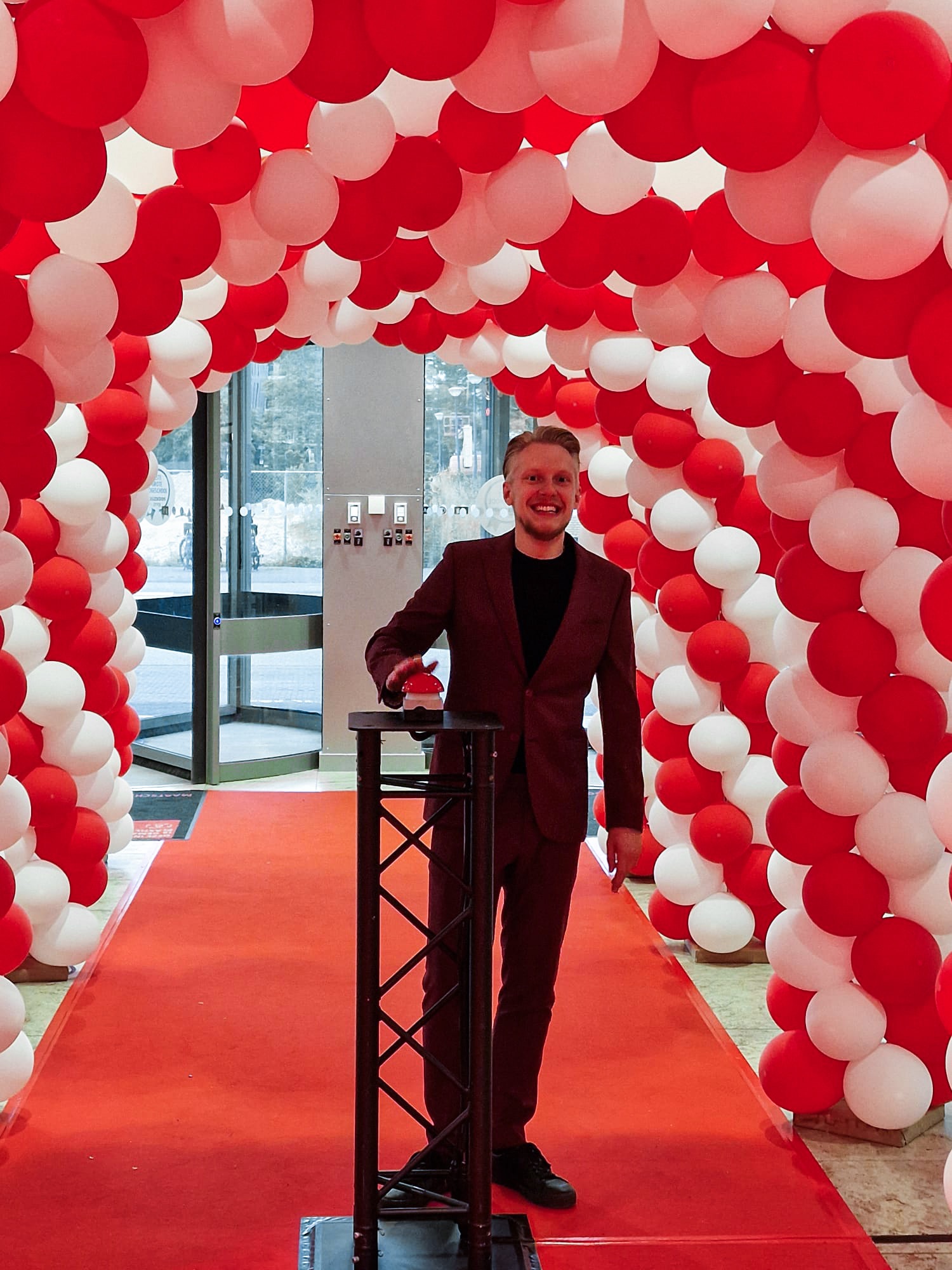 Rick Marcelissen staat onder een boog van rode en witte ballonnen en druk op een grote rode knop.