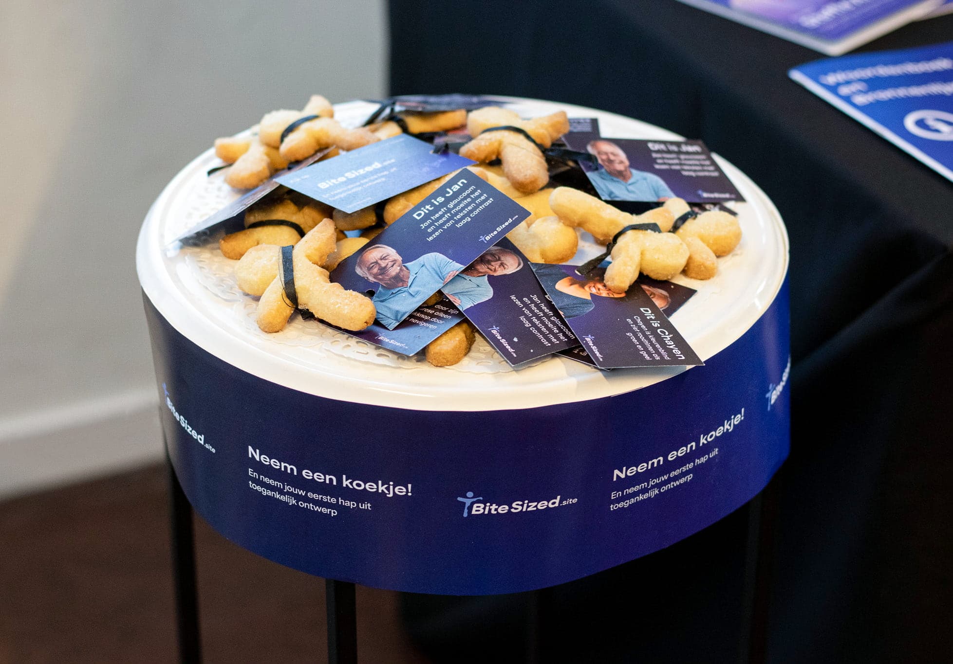 Een schaal met zandkoekjes in de vorm van het logo van BiteSized. Aan elk koekje hangt een kaartje met een voorbeeld een persoon die ontoegankelijkheid ervaart. Op de achterkant staat: 'Ga naar BiteSized.site en neem je eerste hap uit toegankelijk ontwerp.'